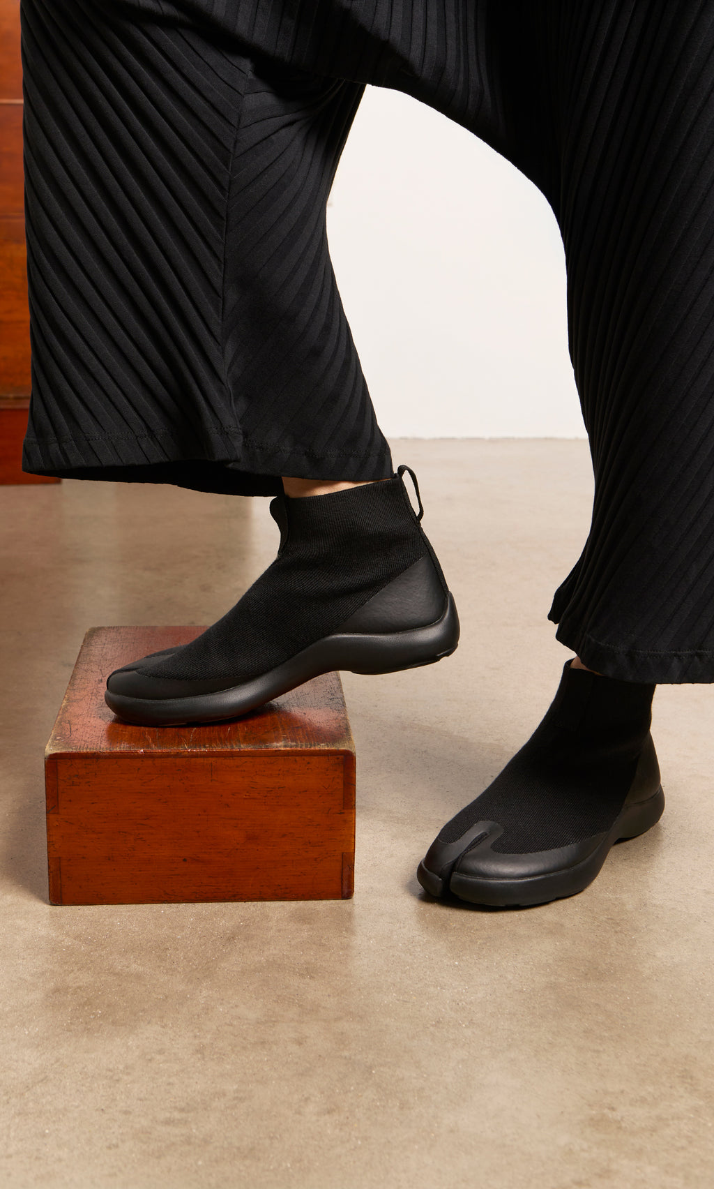 Plümo  Person in pleated black pants and Higher Tabi Boots stands with one foot on a wooden box.