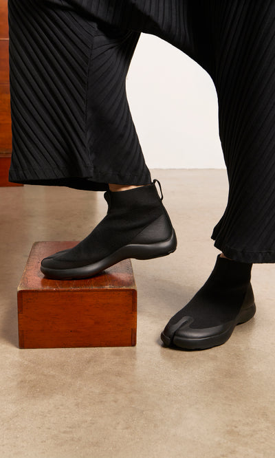 Plümo  Person in pleated black pants and Higher Tabi Boots stands with one foot on a wooden box.