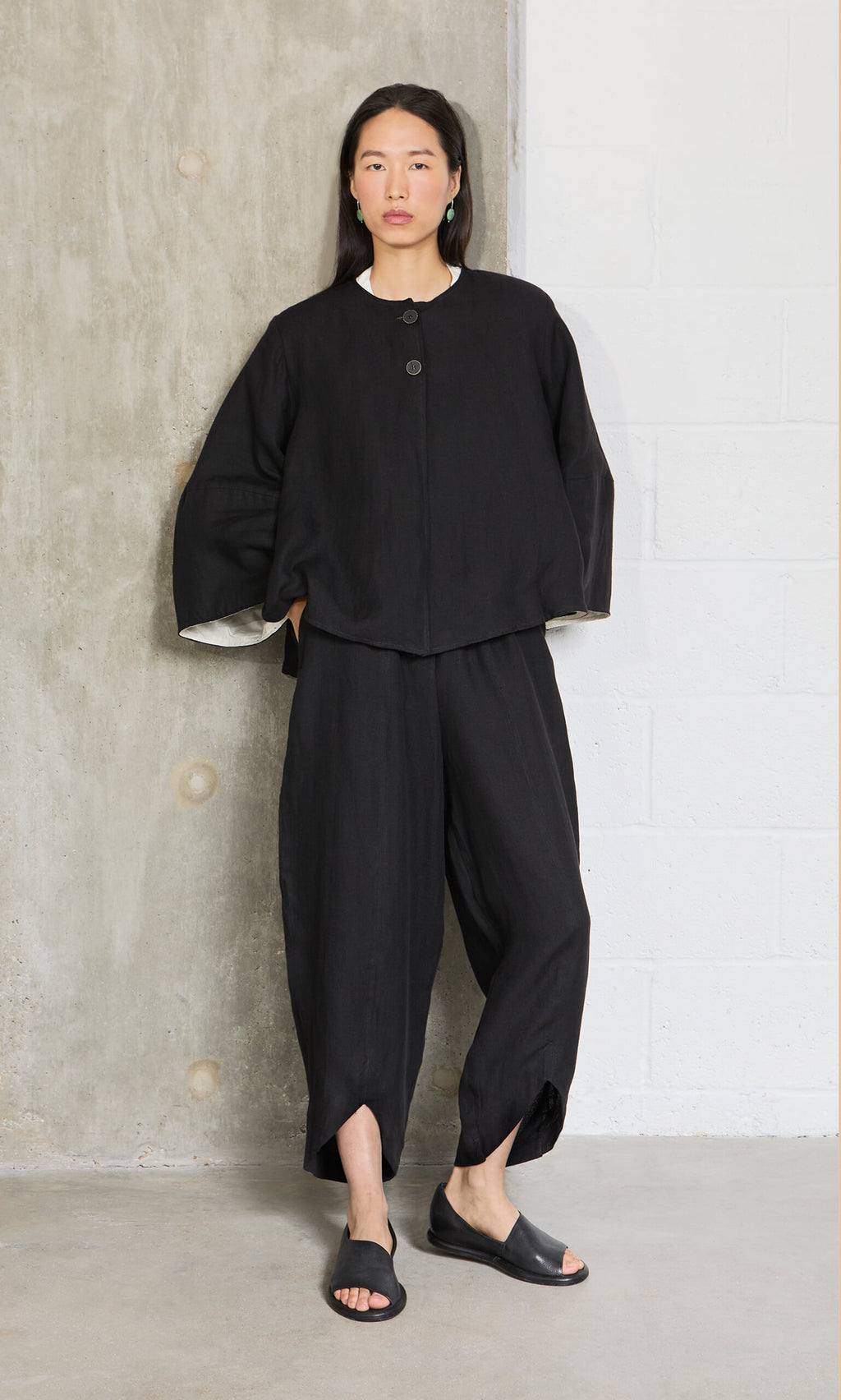 Plümo- A person stands against a concrete wall in Cazalla Pants - Black, looking straight ahead.