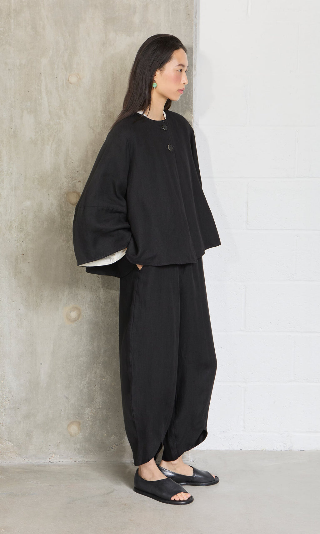 Plümo- A woman in the Holden Jacket, a loose black linen piece with shoulder pads, stands by a concrete wall, looking aside.