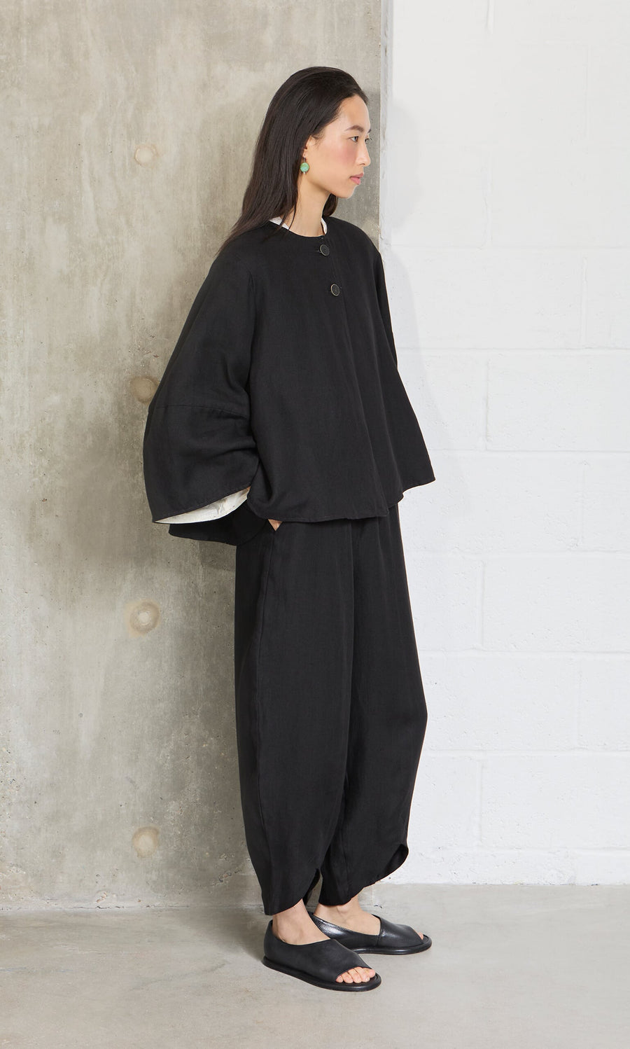 Plümo- A woman in the Holden Jacket, a loose black linen piece with shoulder pads, stands by a concrete wall, looking aside.