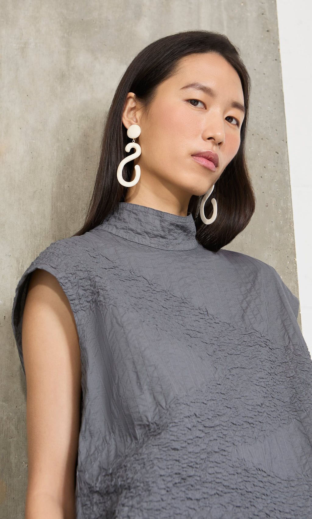 Plümo- Woman with long dark hair wears Horn Clip-On Earrings and a textured gray top, standing by a concrete wall.