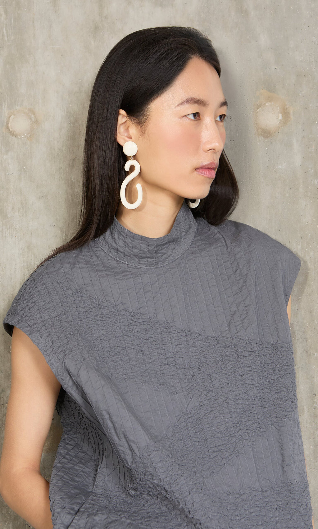 Plümo- Woman with long dark hair in a gray textured top and Horn Clip-On Earrings, standing by a concrete wall.