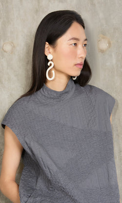 Plümo- Woman with long dark hair in a gray textured top and Horn Clip-On Earrings, standing by a concrete wall.