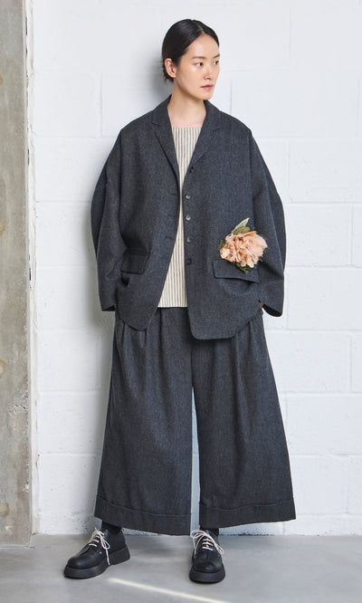 Plümo- Woman in loose dark gray Italian wool Horton Pants, beige top, black shoes, and flower pinned to jacket by a wall.