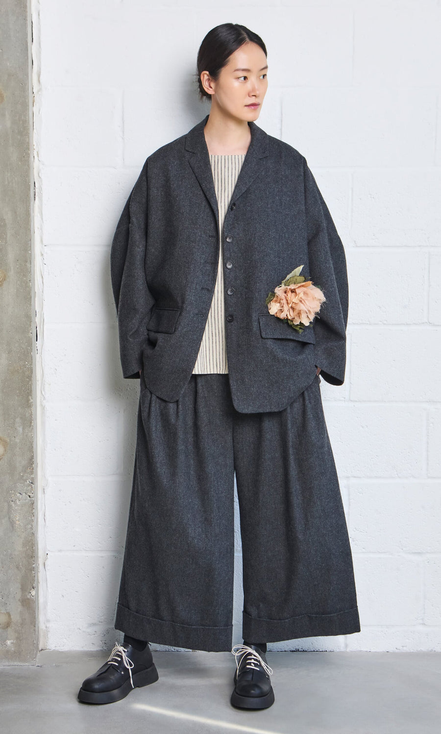 Plümo- Woman in loose dark gray Italian wool Horton Pants, beige top, black shoes, and flower pinned to jacket by a wall.