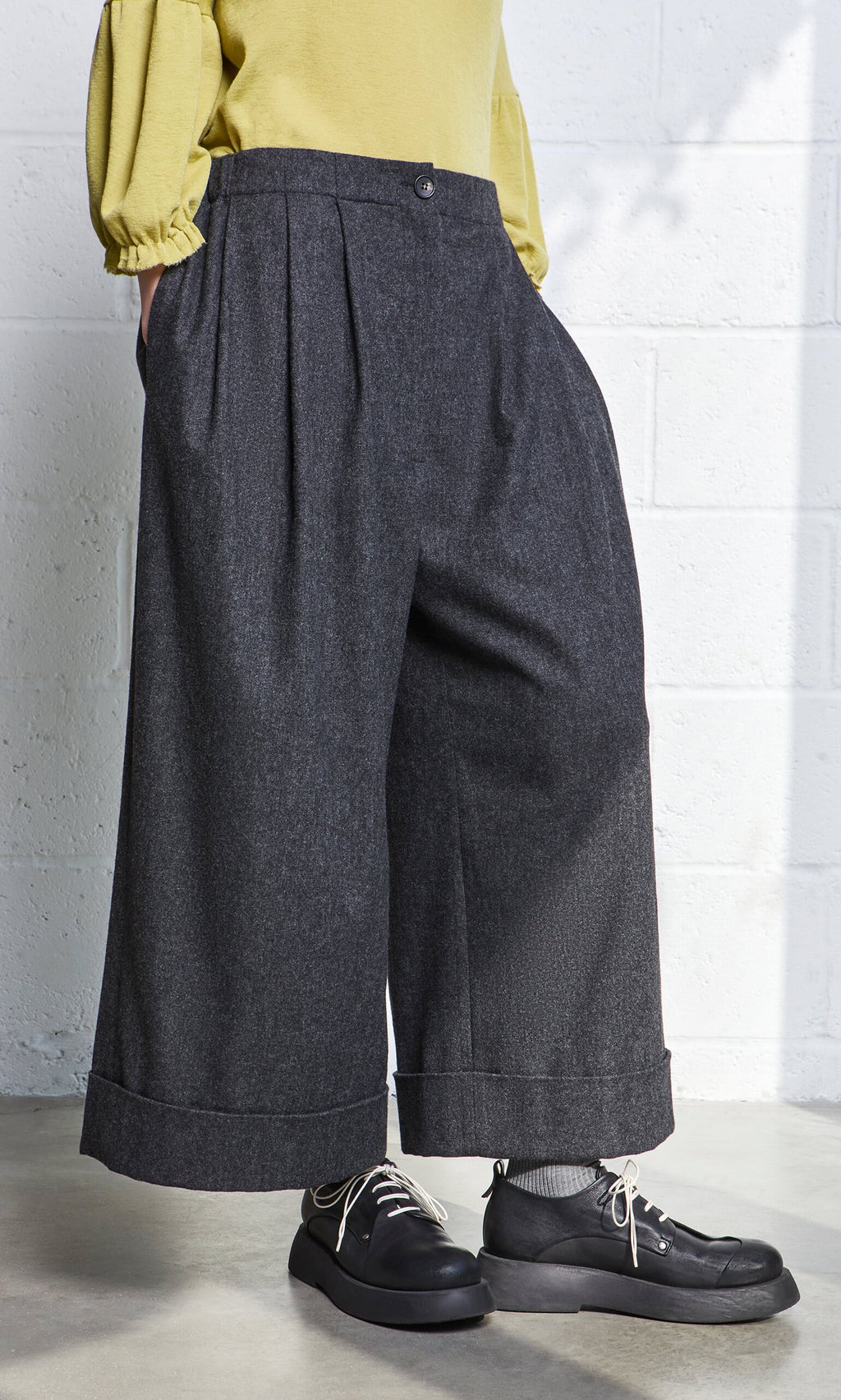 Plümo- Person wearing Horton Pants, black shoes, and a yellow top, standing against a white wall.