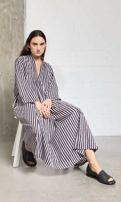 Plümo- Person wearing the Hoyland Dress - Charcoal Stripe sits on a white chair against an industrial-style background.