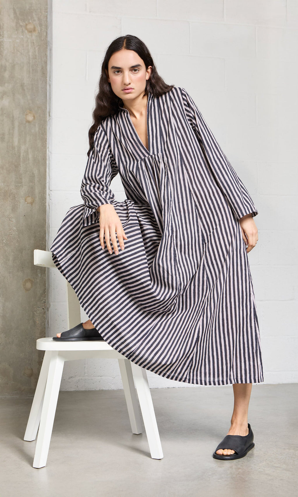 Plümo- Woman in the Hoyland Dress - Charcoal Stripe poses with one knee on a white bench in a minimalistic space.