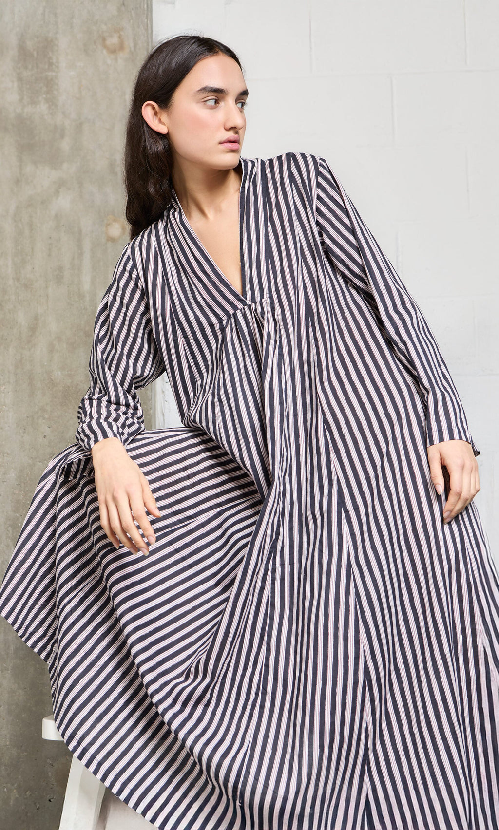 Plümo- Person wearing the Hoyland Dress - Charcoal Stripe, a loose, long-sleeved cotton dress, leaning on a concrete wall.
