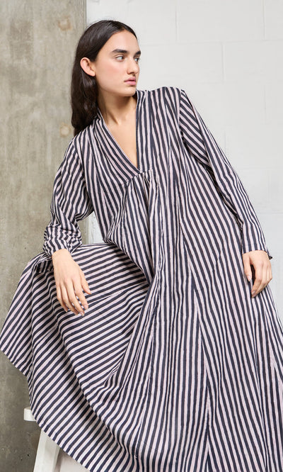 Plümo- Person wearing the Hoyland Dress - Charcoal Stripe, a loose, long-sleeved cotton dress, leaning on a concrete wall.