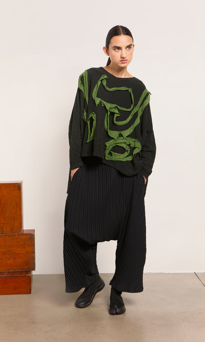 Plümo  Woman in loose black pants and the Isaac Top with an abstract texture stands by a wooden bench.