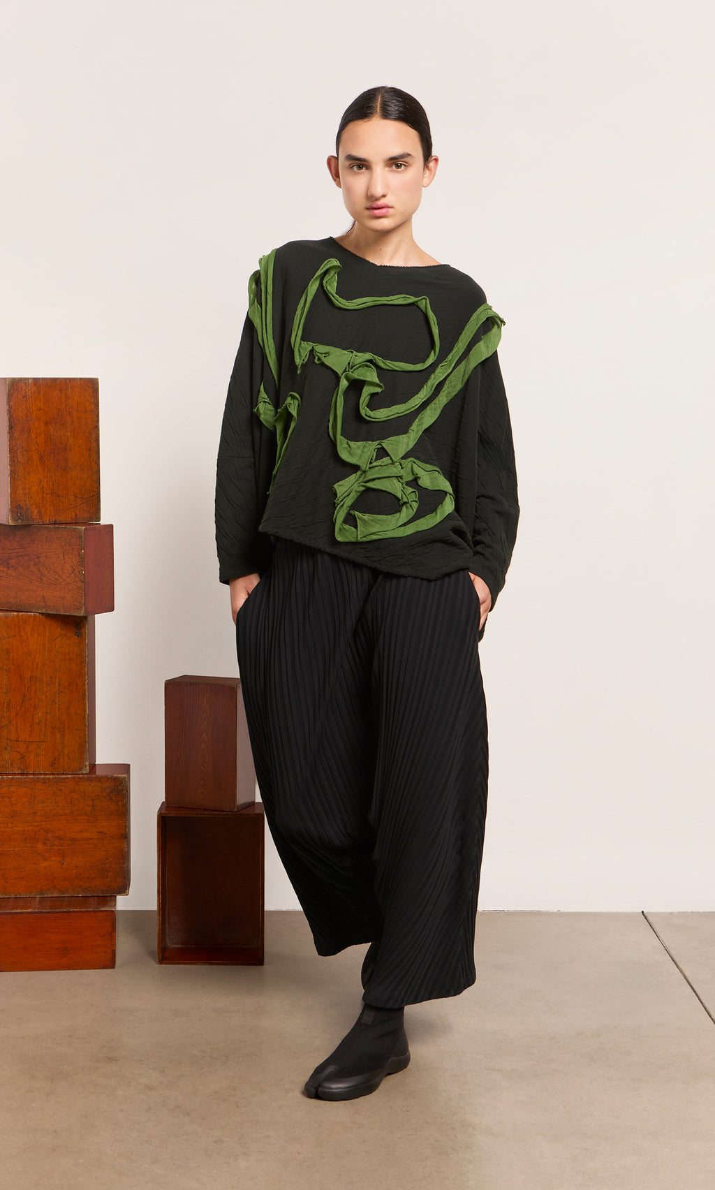 Plümo  Person in ribbed black Acker Pants and a black top with green abstract details, Danish design, stands indoors.