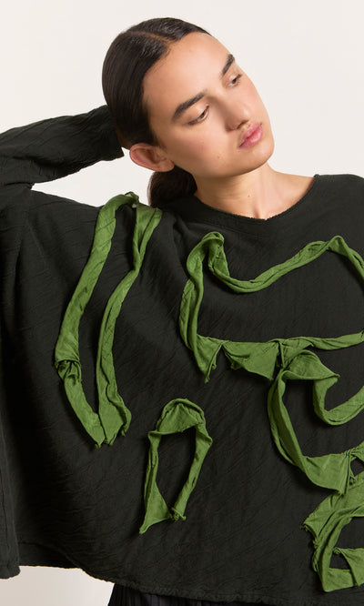 Plümo  Woman in the Isaac Top—a textured black top with green accents and slash neckline—looks to the side, arm raised.