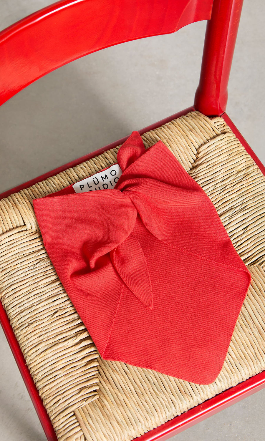 Plümo- The Jona Necktie - Lava rests on a woven straw chair with a red wooden frame.