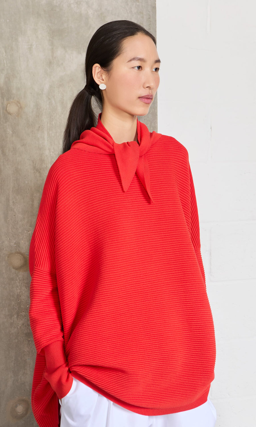 Plümo- Woman with long dark hair wears the Jack Sweater - Lava and white pants, standing against a wall.