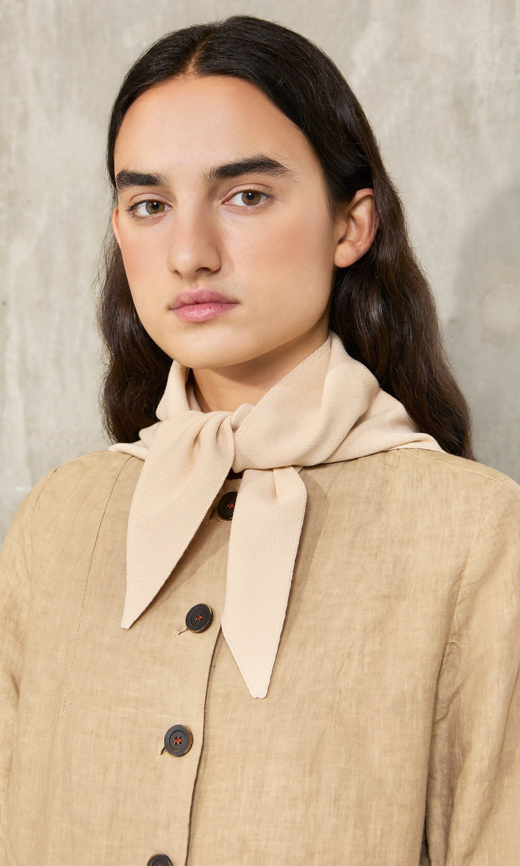 Plümo- Woman with long dark hair wears the Jona Necktie in Silt and a tan top, standing against a gray textured background.