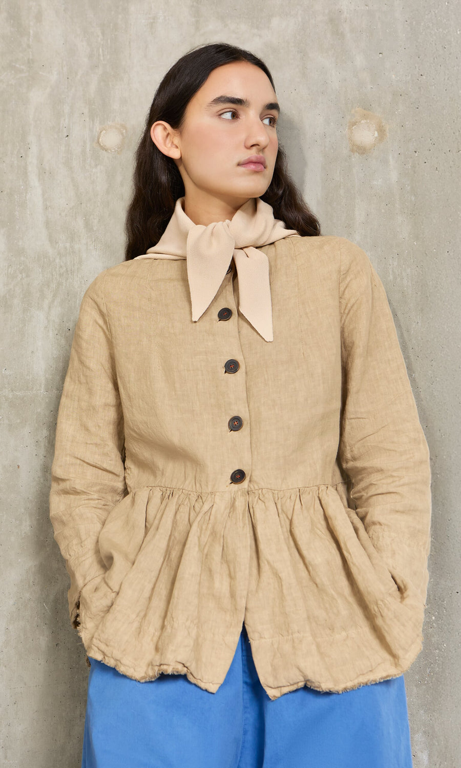Plümo- A person in a tan jacket, Jona Necktie - Silt, and blue pants stands by a textured concrete wall.