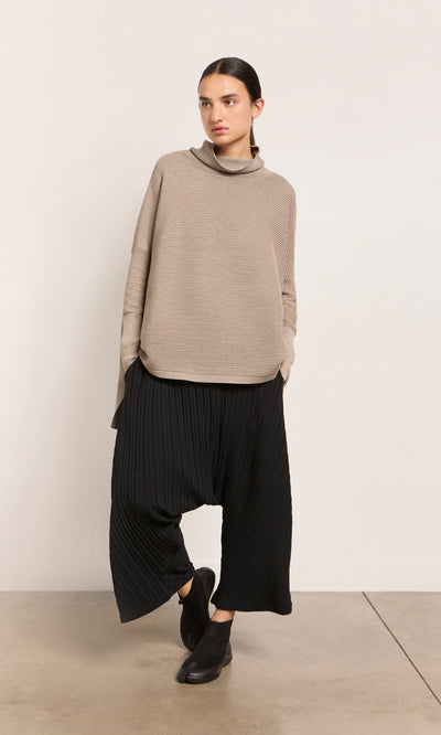 Plümo  Woman in the Jack Rollneck - Miso and wide black pleated pants stands by a plain light wall.
