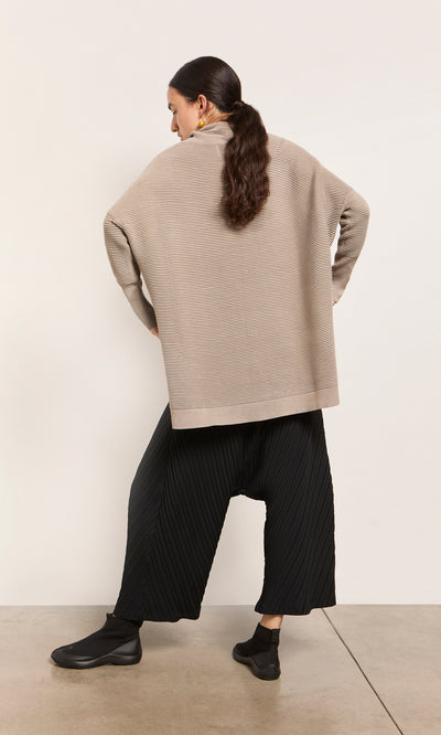Plümo  Person in Jack Rollneck - Miso and black pleated pants, back turned—relaxed look in plant-based fibres.