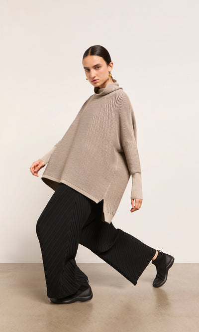 Plümo  Woman in the Jack Rollneck - Miso, beige oversized jumper, with wide black pleated pants, posing against a plain background.