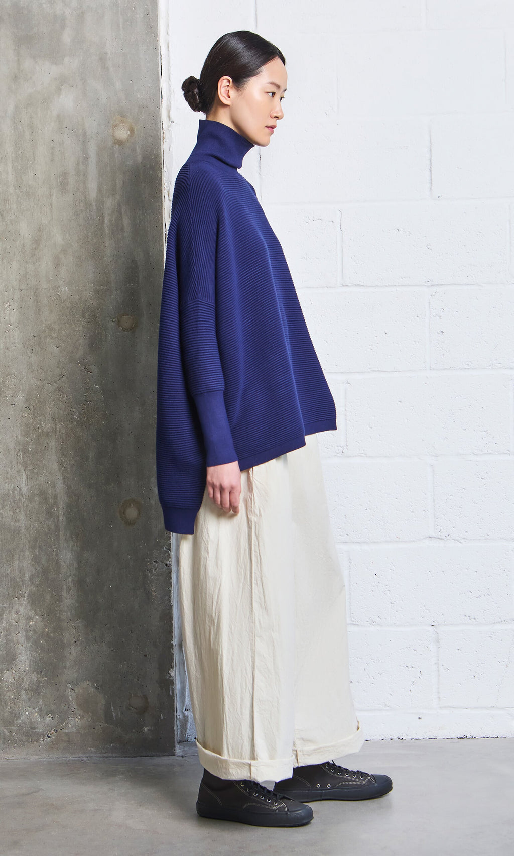 Plümo- Woman in navy turtleneck, Jung Pants, and black shoes stands by a concrete and white brick wall.