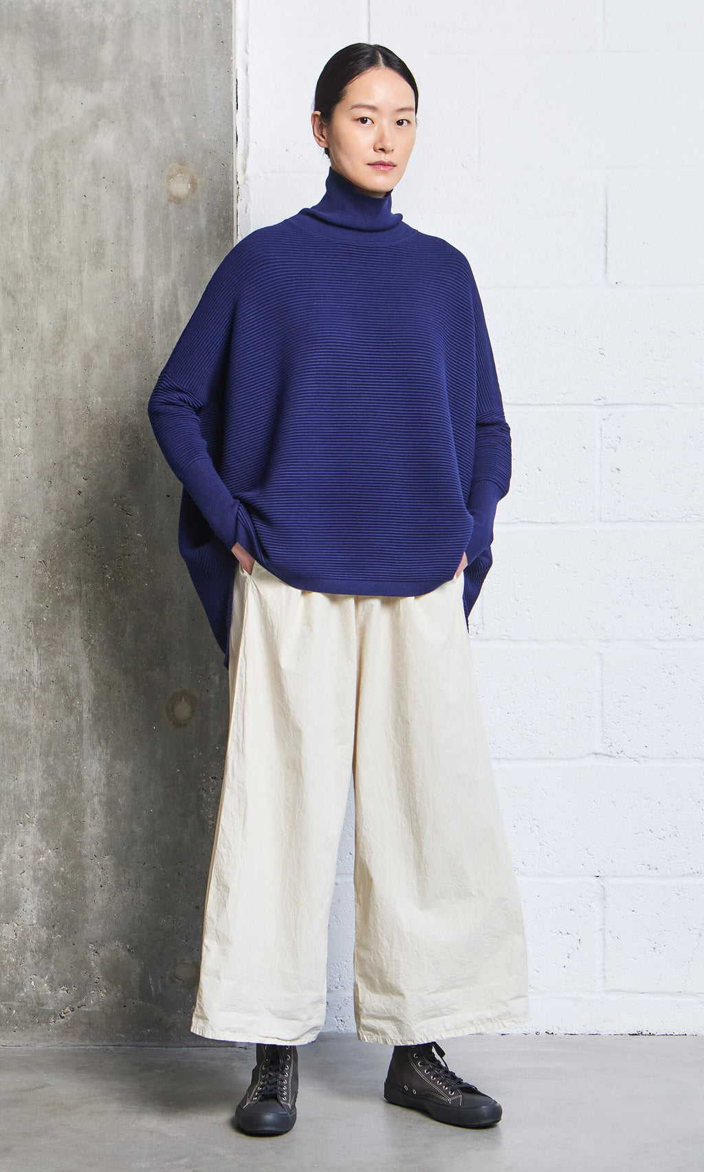 Plümo- Woman in hand-dyed Jung Pants and a blue turtleneck stands against a concrete and white brick wall.
