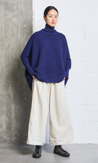 Plümo- Woman in hand-dyed Jung Pants and a blue turtleneck stands against a concrete and white brick wall.
