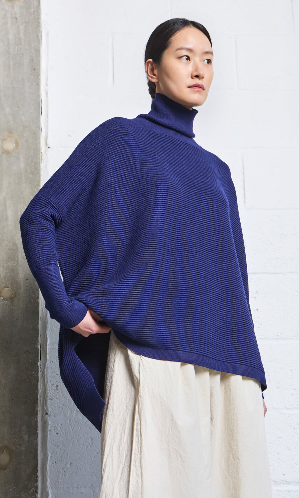 Plümo- Woman wears the Jack Rollneck in navy, paired with a cream skirt, standing against a white wall.