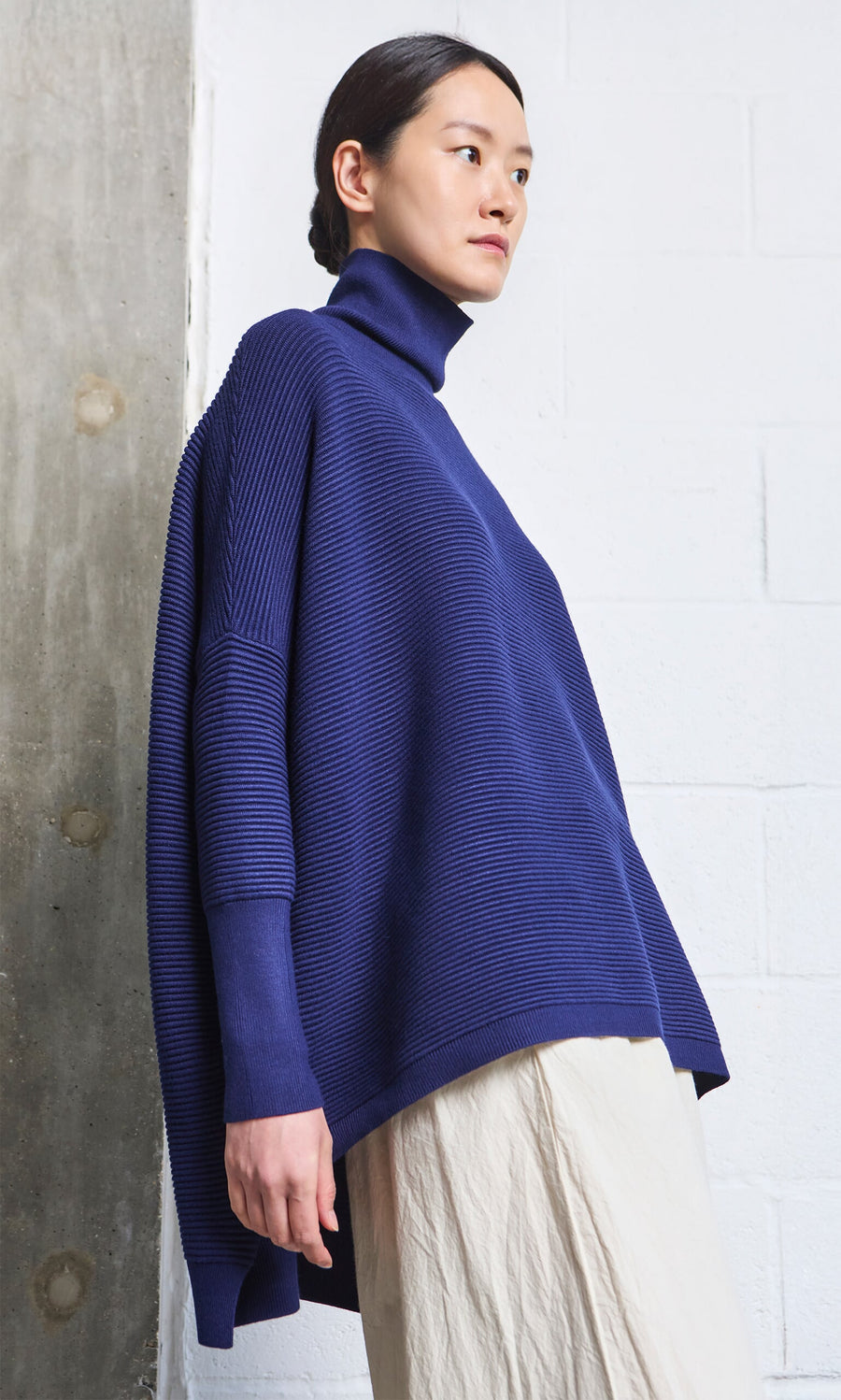 Plümo- Woman wearing the Jack Rollneck - Navy, paired with a light skirt, stands by a white and concrete wall.