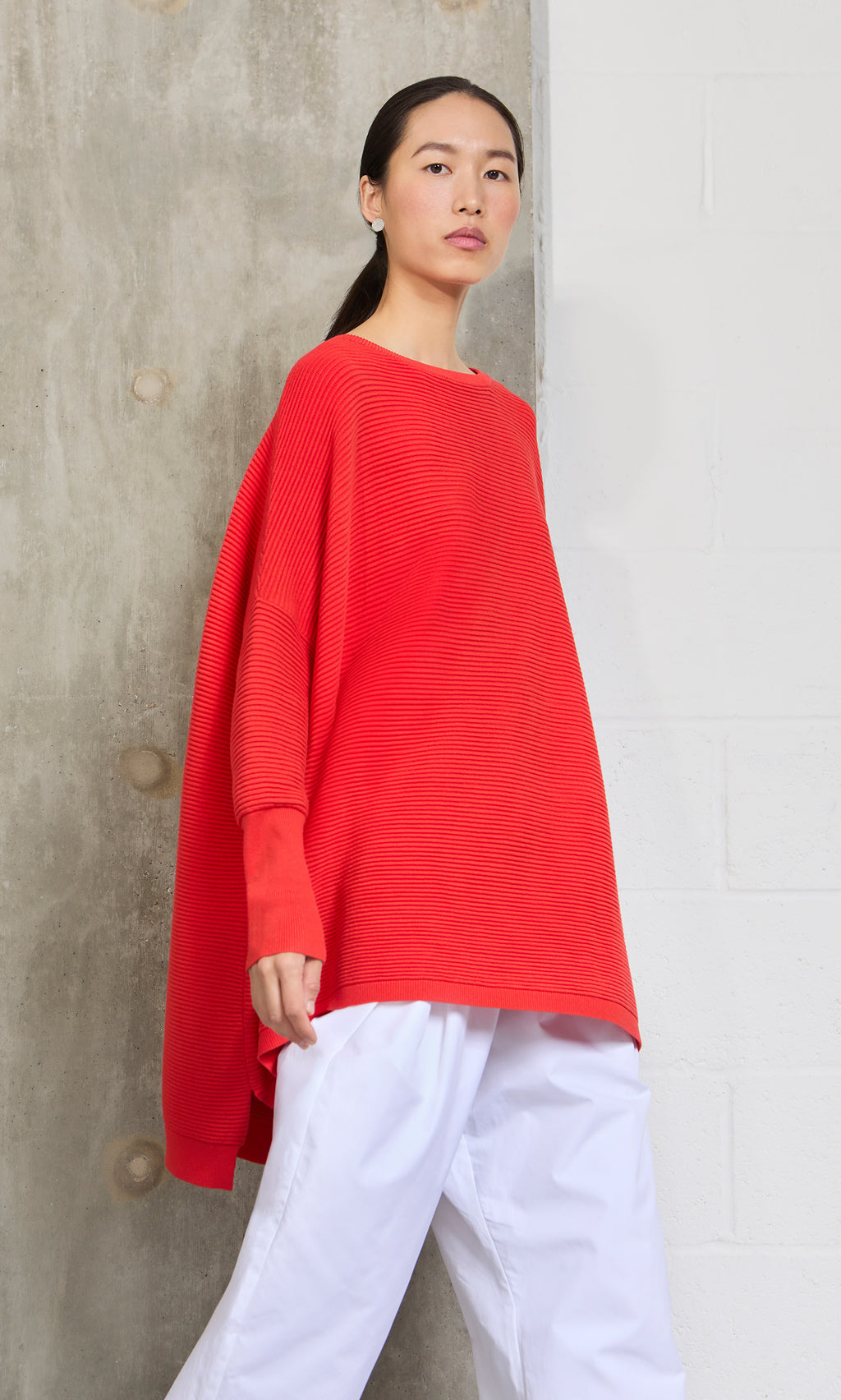 Plümo- Woman wearing the Jack Sweater - Lava, a super soft, oversized red sweater with dropped shoulders and white pants.