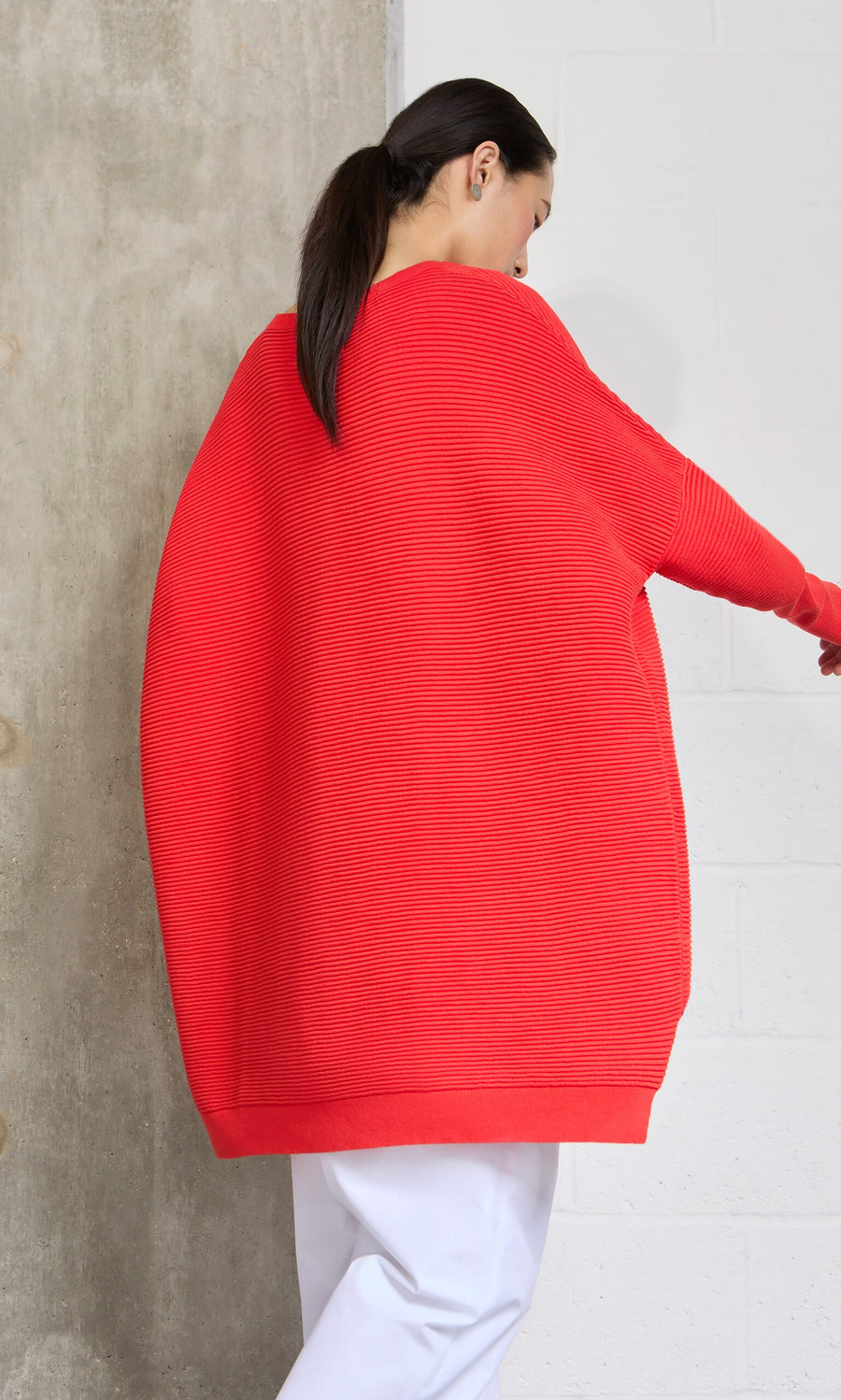 Plümo- Woman with dark hair wears the Jack Sweater - Lava, featuring dropped shoulders and white pants, facing away.