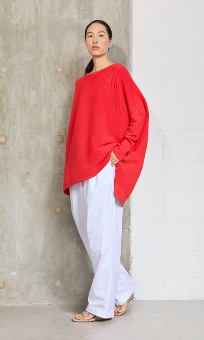 Plümo- Woman in the Jack Sweater - Lava with dropped shoulders and white pants stands confidently by a concrete wall.
