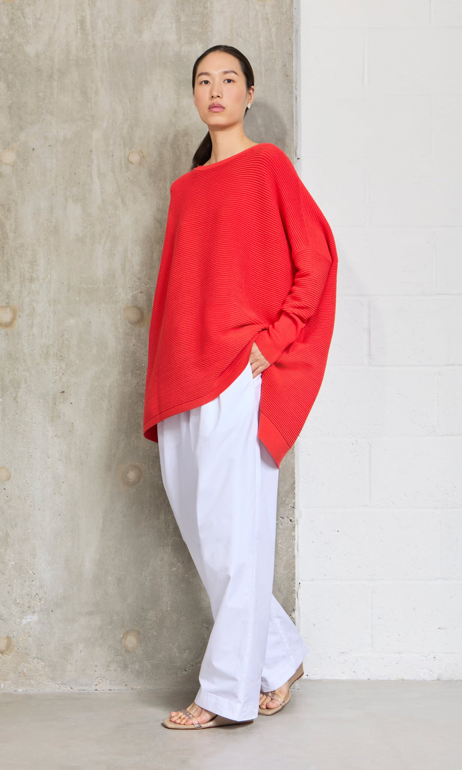 Plümo- Woman in the Jack Sweater - Lava with dropped shoulders and white pants stands confidently by a concrete wall.
