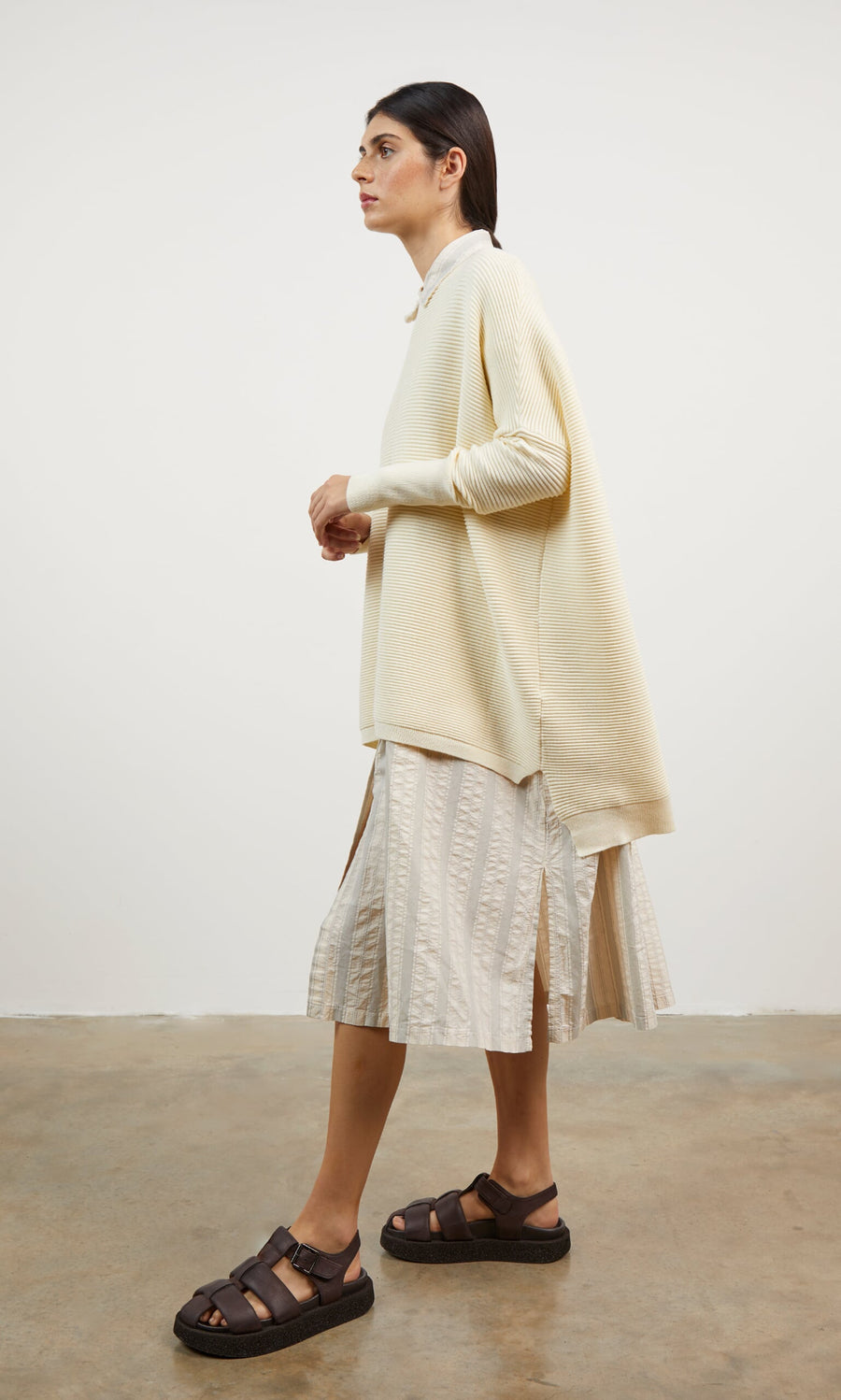 Plümo- Woman wears the Jack Sweater - Cloud, a soft oversized jumper, with a patterned midi skirt and brown sandals.