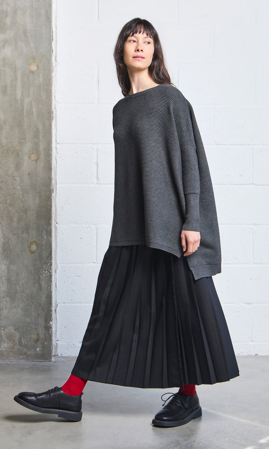 Plümo- Woman in the Jack Sweater - Graphite with a pleated black skirt and red socks stands by a concrete wall.