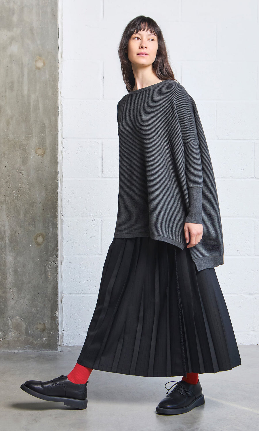Plümo- Woman in an oversized gray sweater, black pleated skirt, and Japanese Red Socks stands indoors.