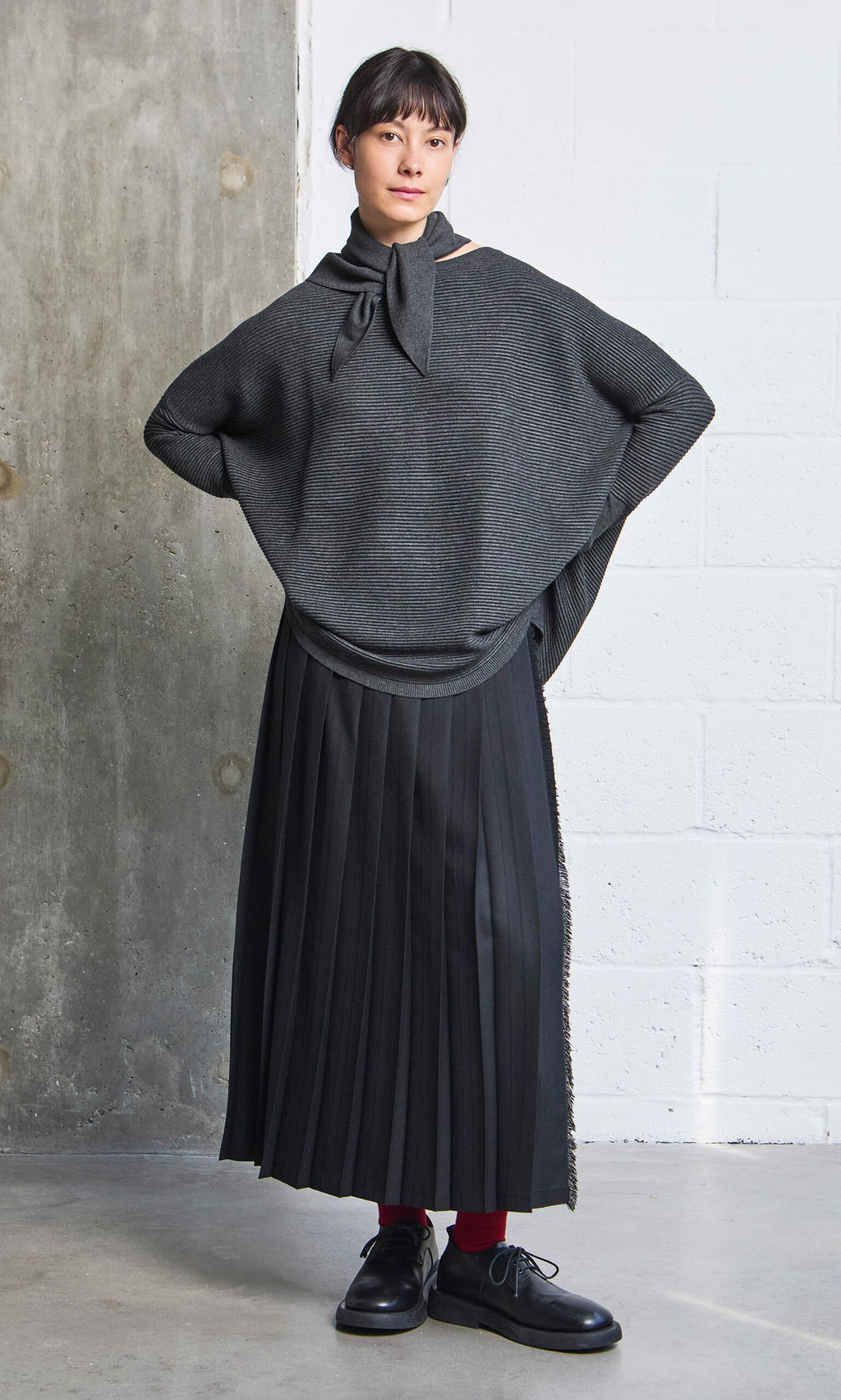 Plümo- Woman in the Jack Sweater - Graphite, black pleated skirt, and red socks stands by a concrete wall.