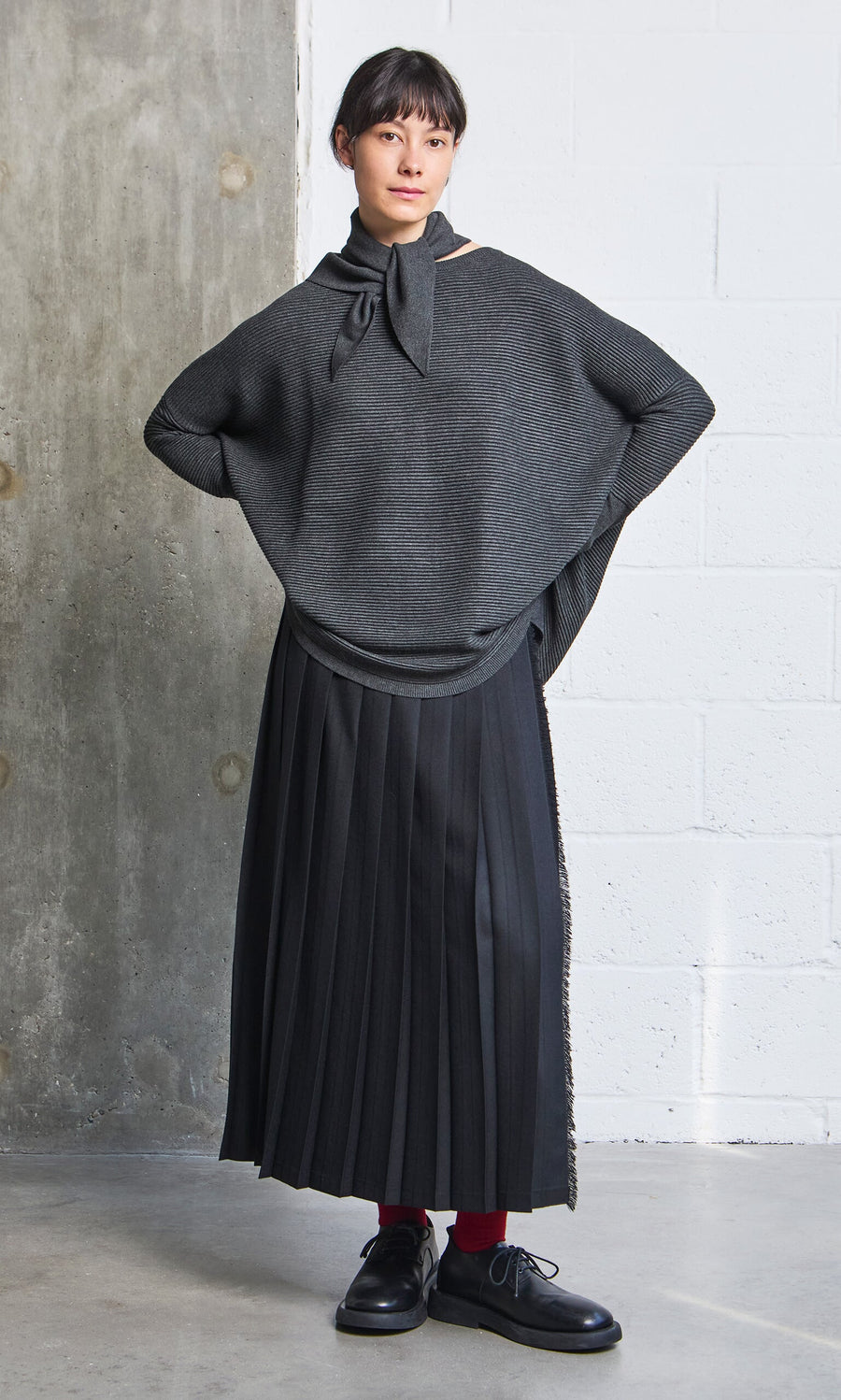 Plümo- Woman in the Jack Sweater - Graphite, black pleated skirt, and red socks stands by a concrete wall.