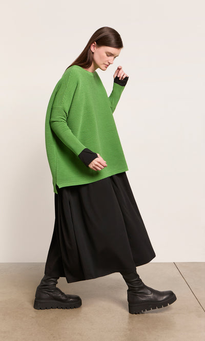 Plümo  A model wears the Jack Sweater in Pickle Green from Plümo Studio, a black midi skirt, and platform boots by a plain wall.