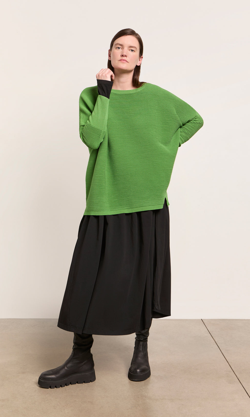 Plümo  Person wears the Jack Sweater in Pickle Green from Plümo Studio, black skirt, and boots against a plain background.