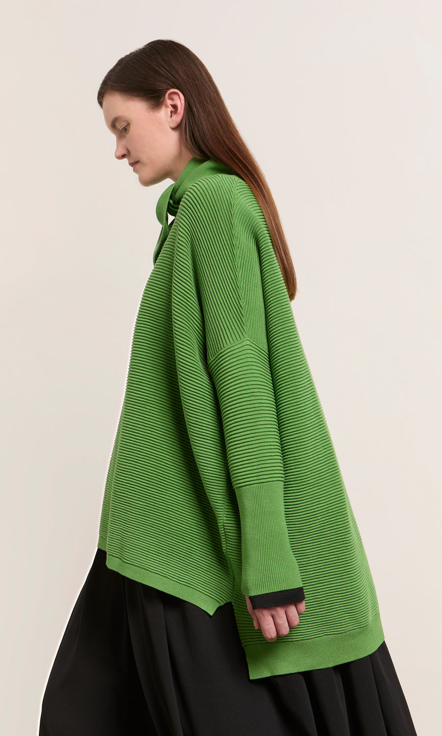 Plümo  A woman wears the Jack Sweater in Pickle Green from Plümo Studio with a black skirt, standing sideways.