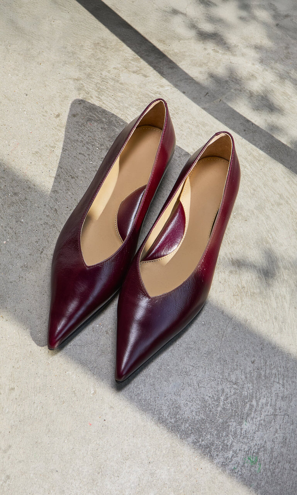 Plümo- Jan Pumps in burgundy patent leather with a low block heel, showcased on a sunlit concrete surface.