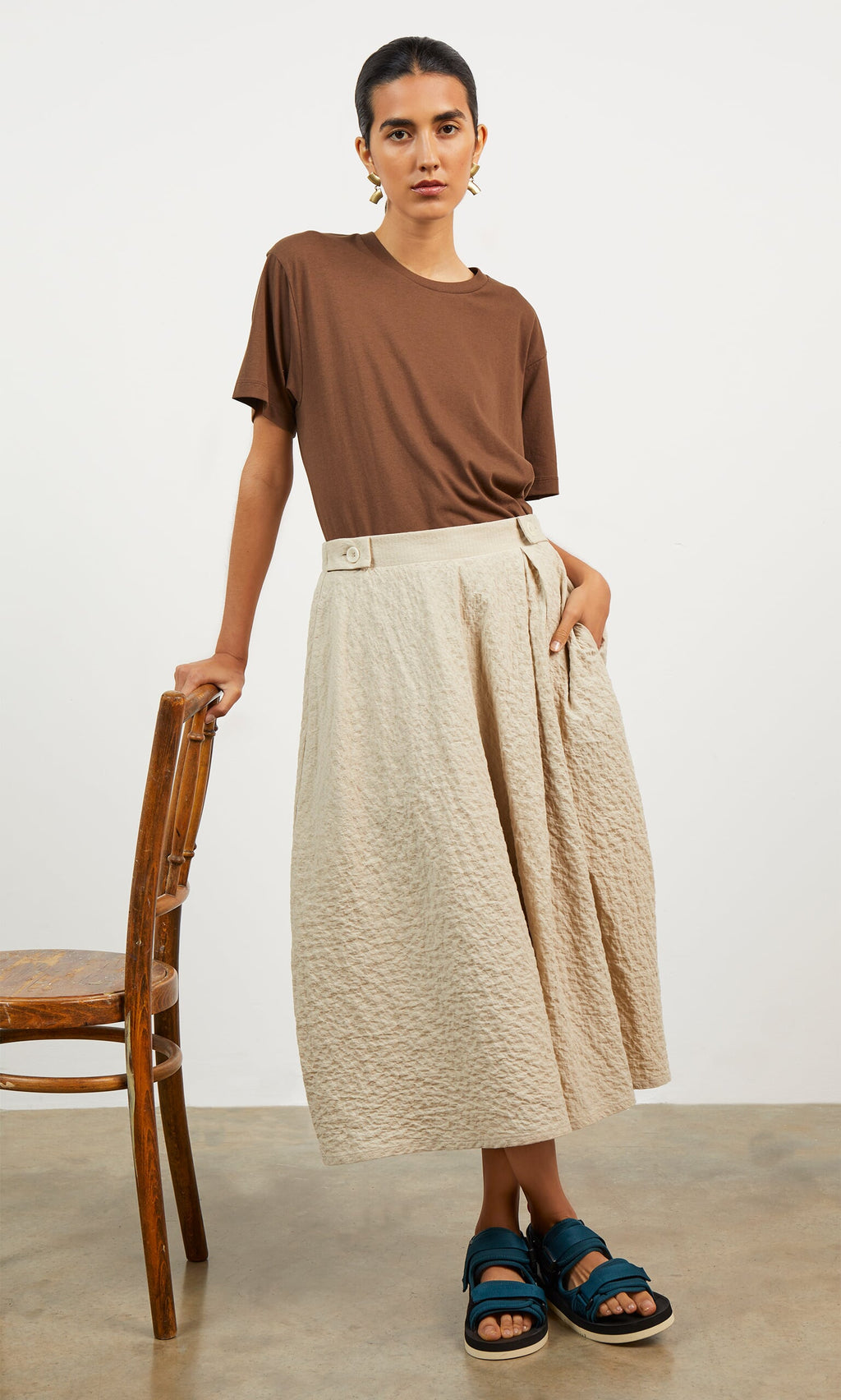 Plümo- A person in a brown t-shirt, Jane Skirt, and blue sandals stands by a wooden chair on a concrete floor.
