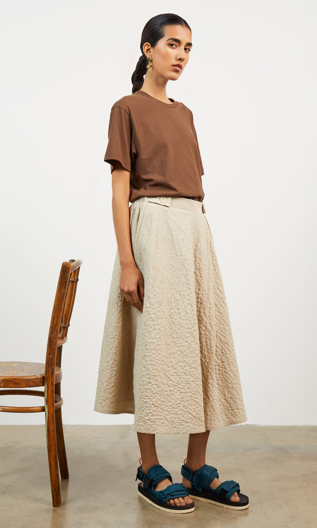Plümo- Woman stands beside a wooden chair in a brown tee, Jane Skirt with asymmetric pleating, and blue sandals.