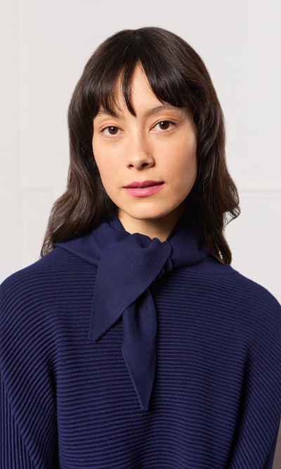 Plümo- Woman with wavy dark hair faces forward in the Jona Necktie - Navy sweater, featuring a tied bow at the neck.