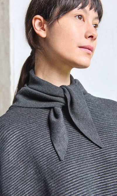 Plümo- Woman in a graphite Jona Necktie and supersoft ribbed gray sweater, gazing thoughtfully to the side against a light background.