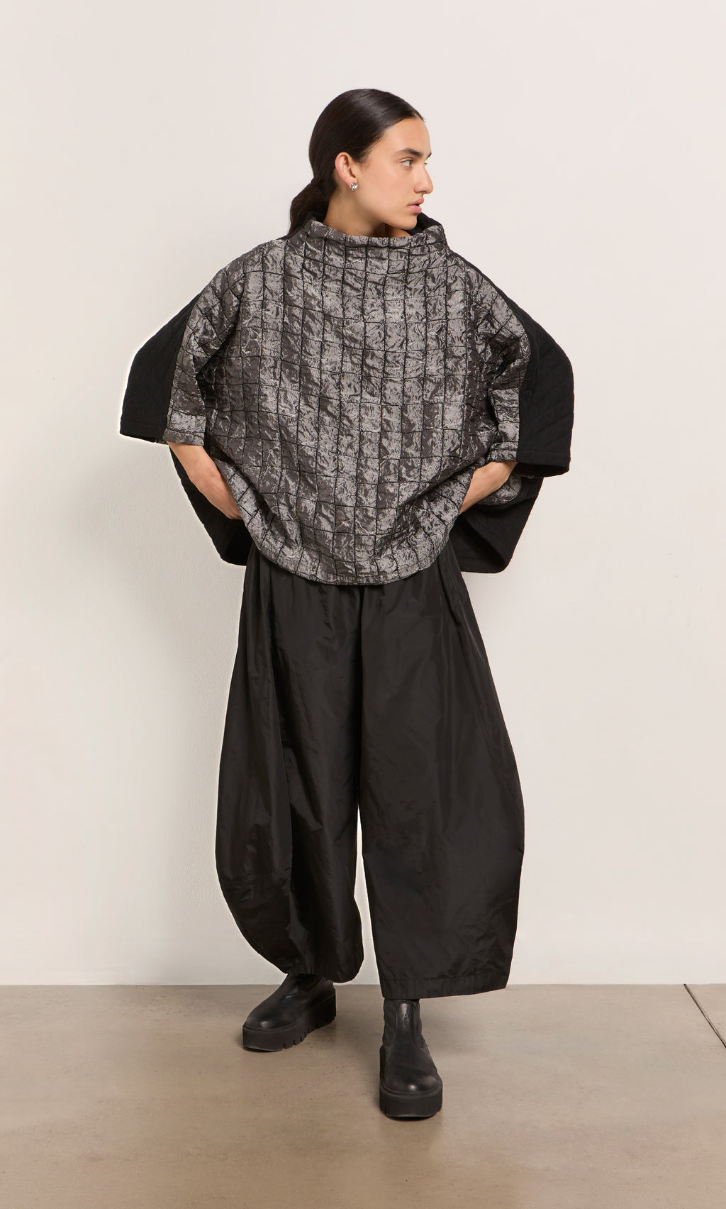 Plümo  Woman in a Jorn Top, wide black pants, and black boots stands against a plain wall, looking to the side.