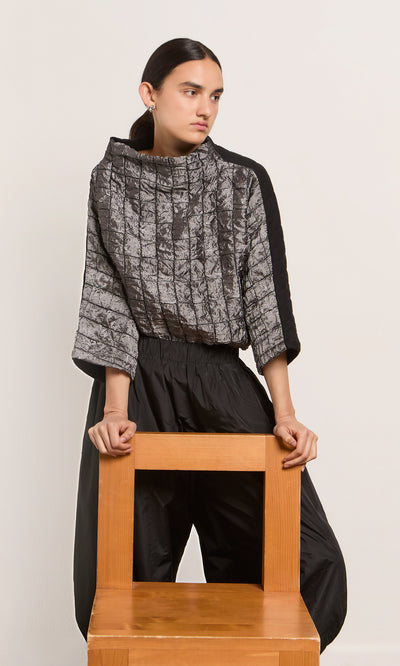 Plümo  Person in an oversized gray Jorn Top and black pants stands behind a wooden chair, looking to the side.