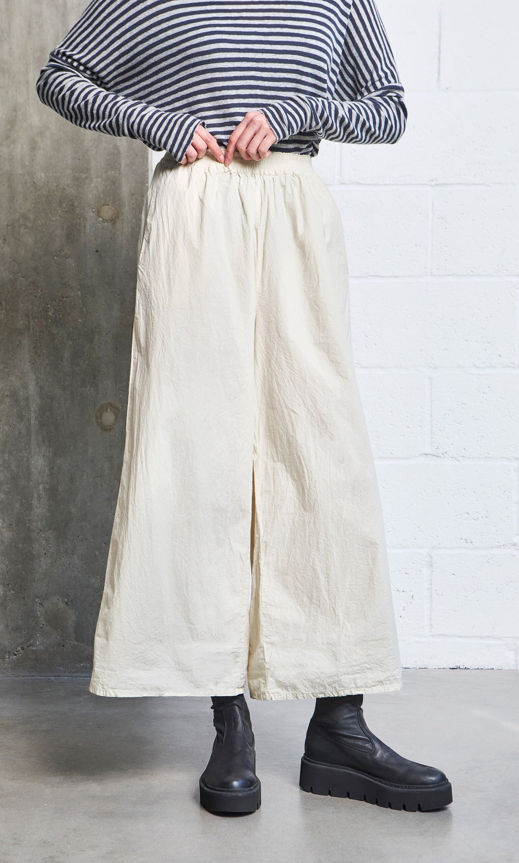 Plümo- Person wearing Jung Pants, a striped long-sleeve shirt, and black boots against a concrete and white wall.
